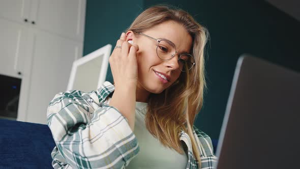 Young Positive Blonde Woman Wearing Eyeglasses Putting in Wireless Earphones and Working Online on