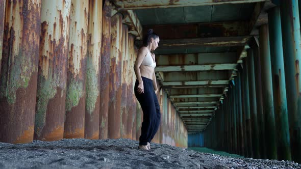 Young Female Dancer is Dancing Alone Under Pier on Pebble Sea Beach Rehearsing