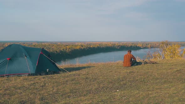 Guy in Orange Shirt Sits at Bonfire By Blue Tent in Morning