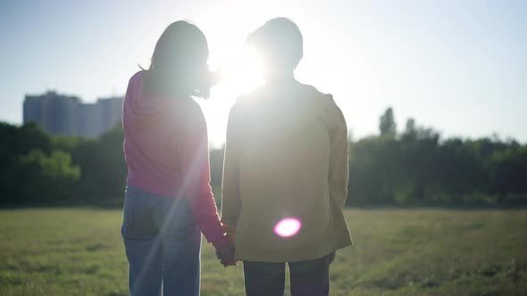 Back View Relaxed Loving Teenage Couple Holding Hands Standing in Sunbeam in Summer Park