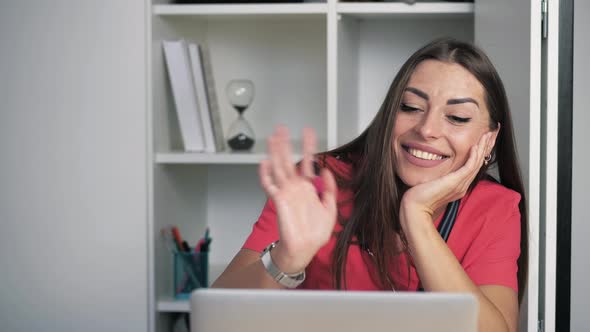 Young Caucasian Woman Doctor in Coral Gown Sittting at Table in Cabinet and Videochatting on Laptop