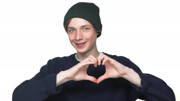 Portrait of Smiling Caucasian Youngster Wearing Hat Showing Heart Shape with Fingers Being Shy Over