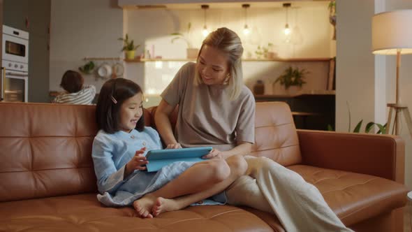 Loving Asian Mom and Little Daughter Sit Together on Sofa at Home With Tablet Device Draw With