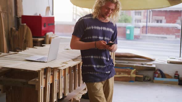 Caucasian male surfboard maker standing in his studio and using his smartphone