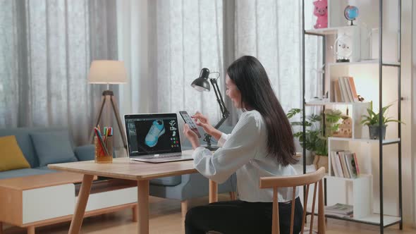 Asian Female Footwear Designer Looking At Photos On Smartphone While Designing Shoe On A Laptop
