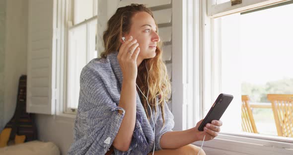 Happy caucasian woman wearing earphones sitting using smartphone in sunny cottage living room