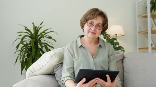 Beautiful Old Lady Looking in Camera Sitting in the Living Room with Tablet