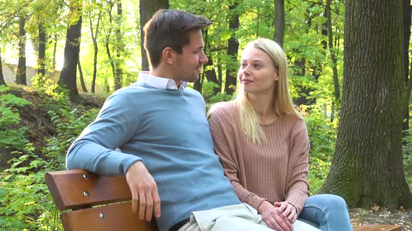 A Young Couple Sits on a Bench and Wave at the Camera with a Smile in a Park on a Sunny Day