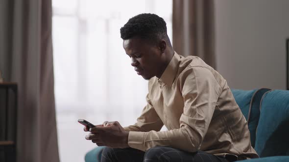 Young Man is Sitting in Apartment and Using Smartphone Afroamerican Guy is Surfing Internet By
