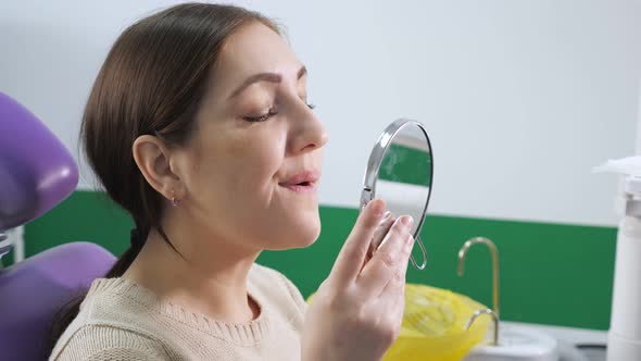 Young Woman in Dental Chair Examines Her Teeth in the Mirror After Treatment