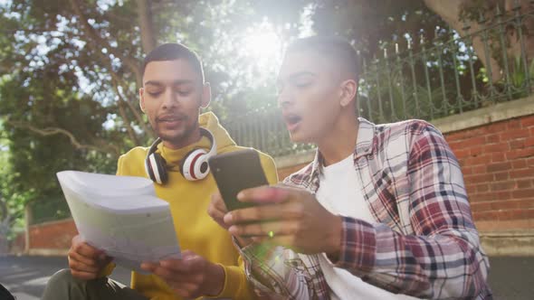 Two happy mixed race male friends sitting, using smartphone and map