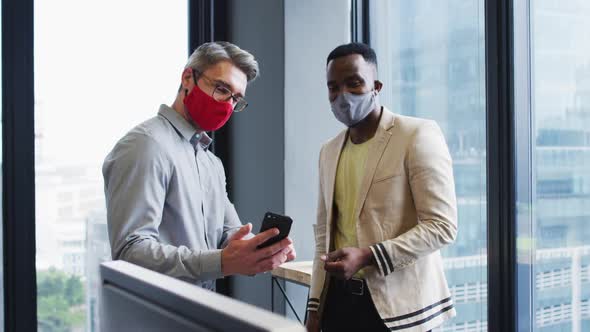 Diverse male office colleagues wearing face masks discussing over smartphone at modern office