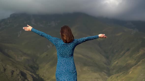 A Young Woman Stands in a Blue Dress and Raises Her Hands Up Looking at the Mountains.