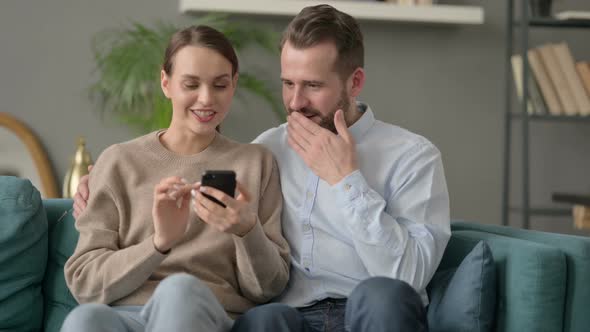 Portrait of Couple Celebrating Success on Smartphone Sitting on Sofa