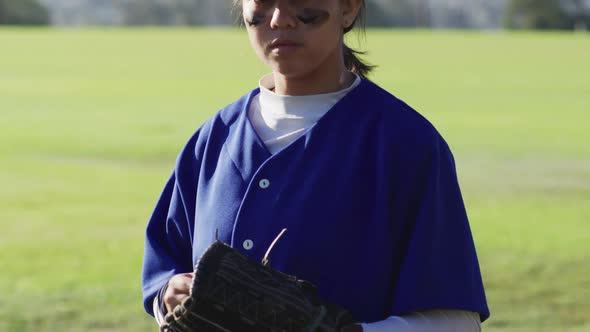 Portrait of mixed race female baseball player wearing eye black, throwing ball into glove
