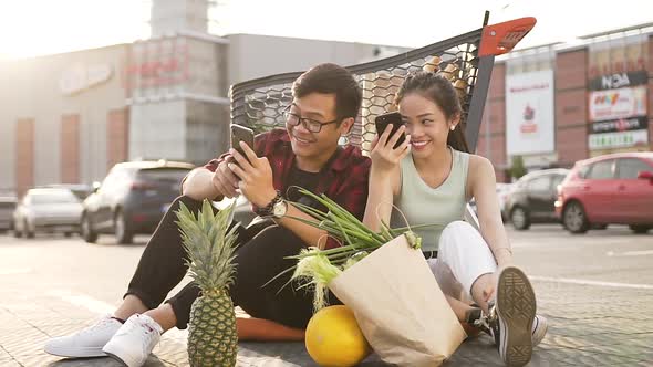 Asian Man and Woman Sitting on the Asphalt Near Market Trolley