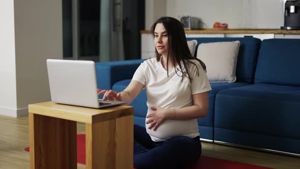 A Pregnant Woman in Home Clothes is Sitting on the Floor and Using a Laptop