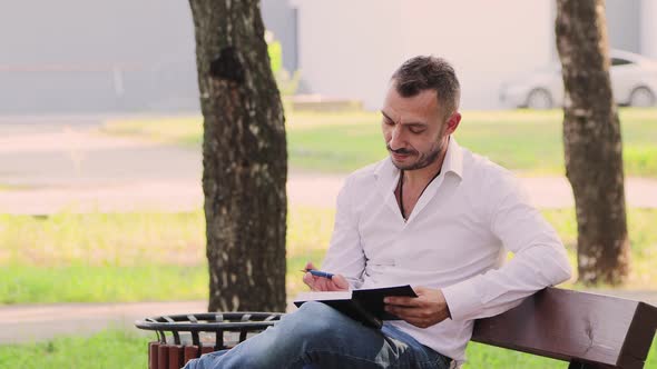 A Business Man in a White Shirt Writes Down Thoughts in a Notebook on a Bench