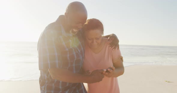 Senior african american couple holding hands and using smartphone on sunny beach