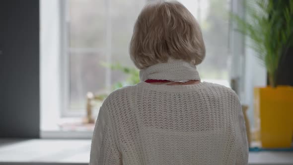 Back View Lonely Senior Caucasian Woman with Grey Hair Looking Out the Window Standing Indoors