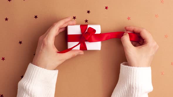 Human hands in white sweater tied and untied a white paper gift with a red satin ribbon bow on brown alt