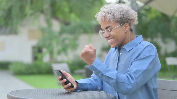 Young African Woman Celebrating Success on Smartphone in Outdoor Cafe