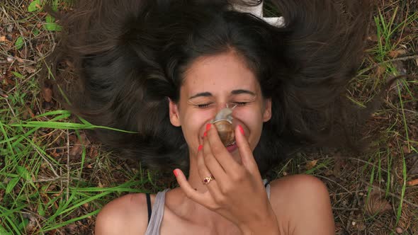 A pretty brunette woman laying in the grass in a forest while placing a snail on her nose and acting