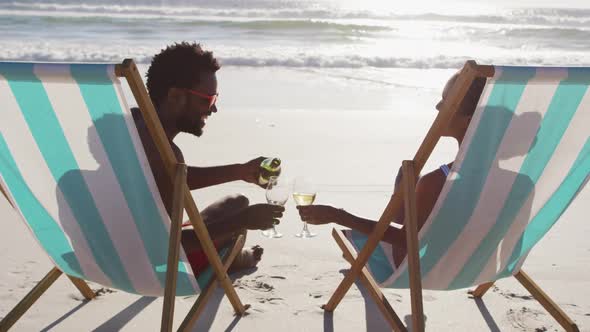 African american couple drinking wine together sitting on deck chairs at the beach