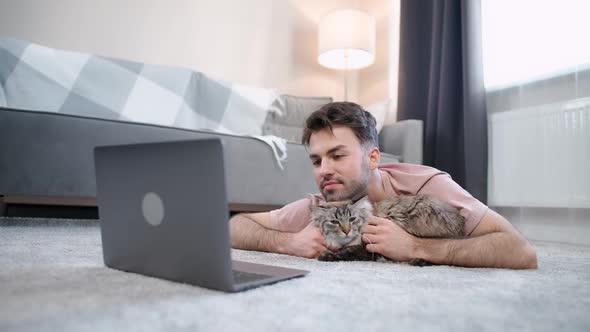 Young Man Lies on the Floor in the Living Room with a Cat and Works at a Laptop Selfisolation at