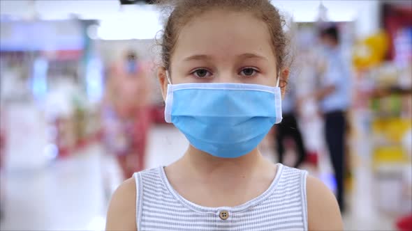 Cute Little Girl From an Epidemic of Coronaviruses or Viruses Looks at the Camera Amid Masked People