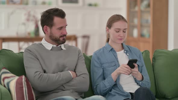 Woman Busy with Smartphone Sitting Next To Angry Man on Sofa