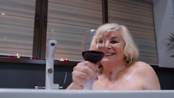 Senior Woman Grandmother Is Taking Foamy Bath, Drinking Red Wine in Luxury Bathroom with Candles