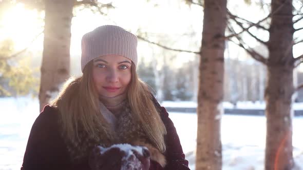 A Young Girl Blows a Pile with Snow From Her Hands Against the Background of Snow and Sun