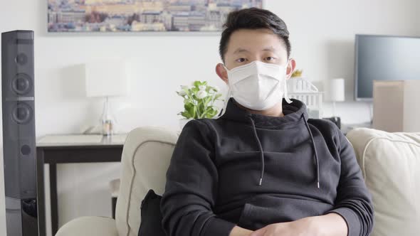 A Young Asian Man in a Face Mask Looks Seriously at the Camera As He Sits on a Couch at Home