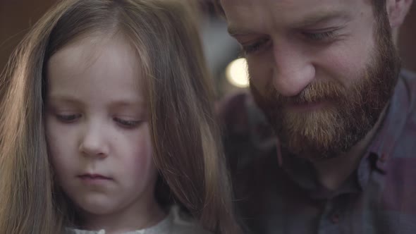 Close-up Portrait of Bearded Man Sitting at the Table in the Kitchen with His Daughter Counting