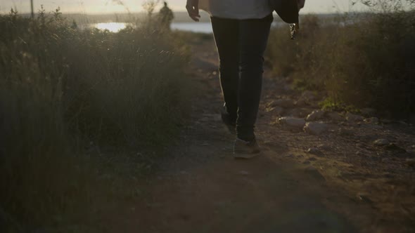 Man Holding a Guitar in a Hand and Walking on a Countryside Trail Against Sunset  Back View