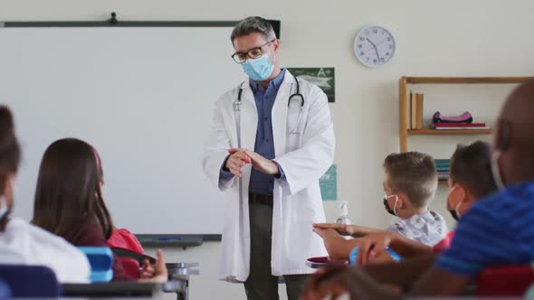 Diverse teacher disinfecting hands in classroom, with schoolchildren sitting wearing face masks