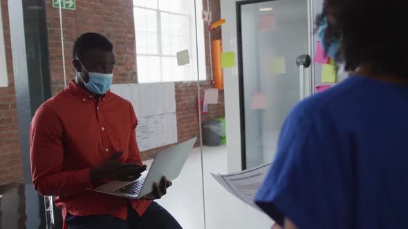 African american businessman with laptop discussing memo notes with colleague wearing face mask