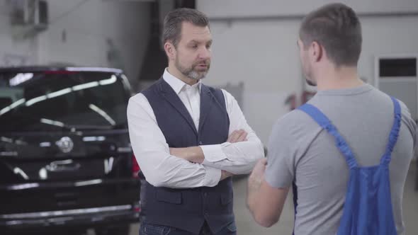 Portrait of Unsatisfied Adult Caucasian Man Talking with Auto Mechanic in Repair Shop. Handsome