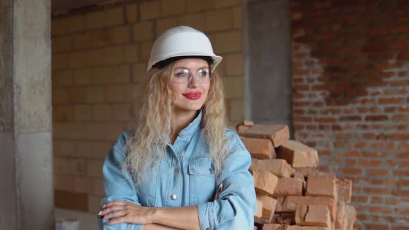 A Young Woman in a Work Helmet and Denim Clothes and Goggles Stands at the Construction Site