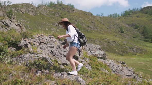 Attractive Brunette Girl in a Hat with a Backpack Travels in the Mountains. Woman in a White Shirt