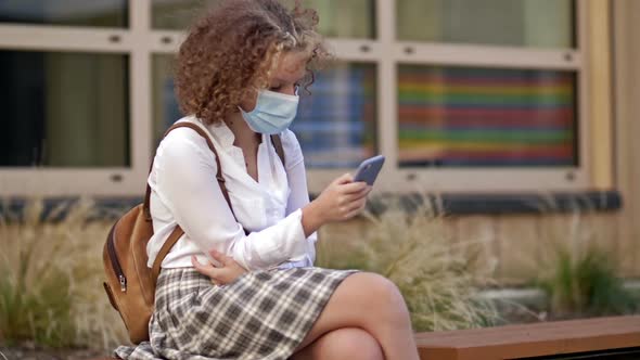 Teenage Schoolgirl in Protective Mask Sits on a Bench Near the School Building with a Smartphone in