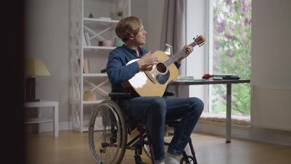 Happy Talented Songwriter Playing Guitar Singing Sitting in Wheelchair at Home in Living Room