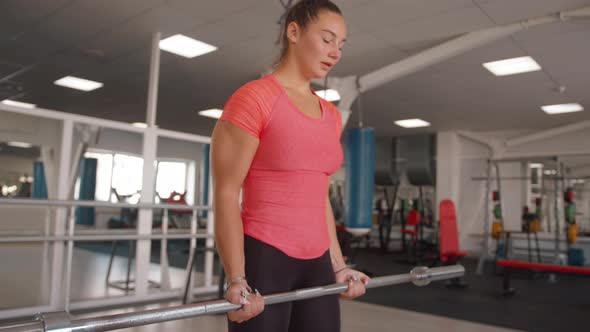 A Young Girl Training in the Gym and Performs a Barbell Lift for Biceps While Standing