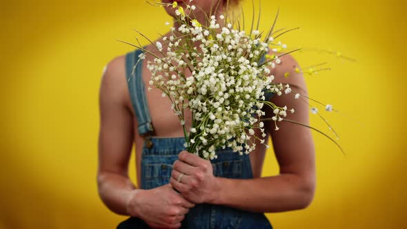 Unrecognizable Young Queer Man in Denim Overalls Posing with White Flowers at Yellow Background