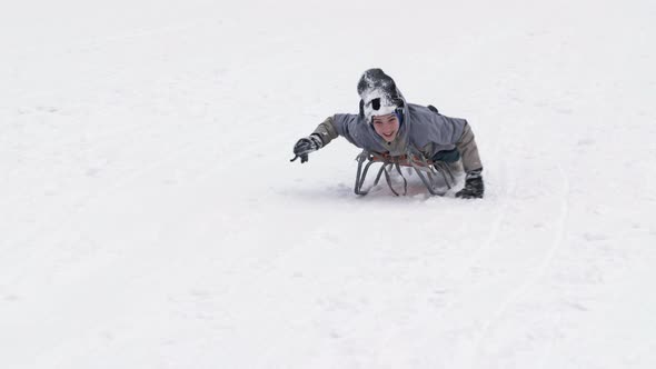 Cheerful Boy in a Ski Suit Slides Down the Slope on a Sled Helping Himself with His Hands