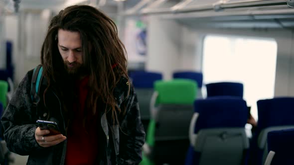 Portrait of a Bearded Hippie Man with Dreadlocks and Big Ones Standing in a Train Car Typing a Text