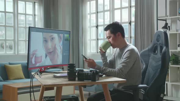 Asian Cameraman Using Mobile Phone And Drink Coffee While Using Computer For Editing Photo At Home