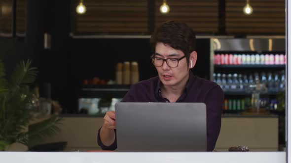 Asian man with laptop wearing wireless earphones drinking coffee while sitting at a cafe