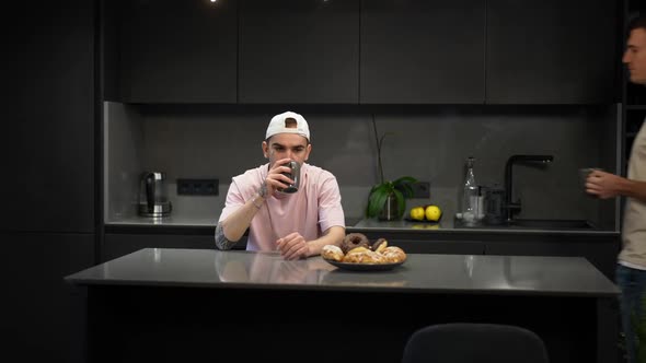 Portrait of Confident Happy Gay Man Drinking Coffee in Kitchen As Partner Entering Hugging Boyfriend